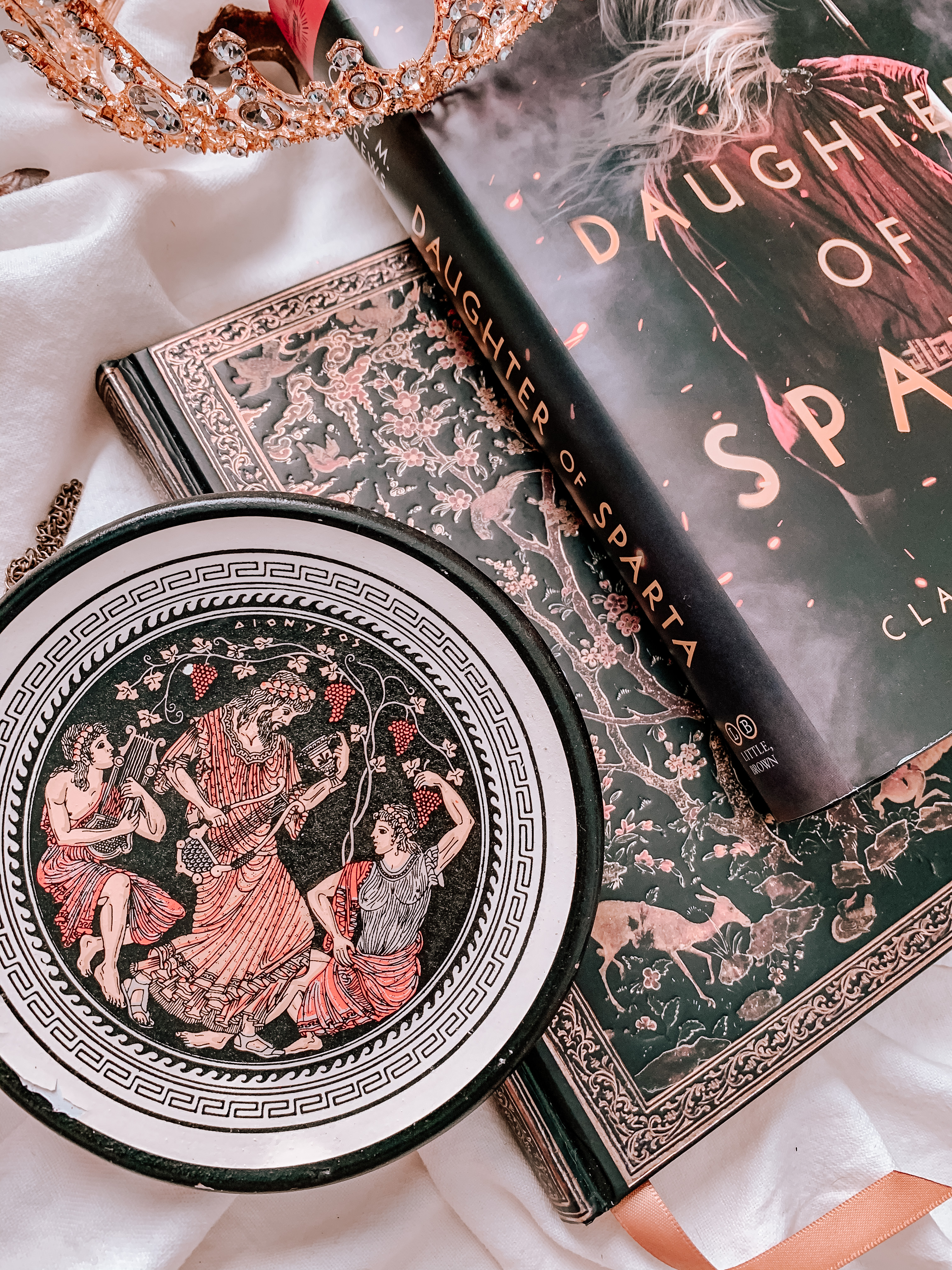 Close-up of a Greek dish hand painted with Dionysus on it, next to a notebook and the hardcover edition of Daughter of Sparta.
