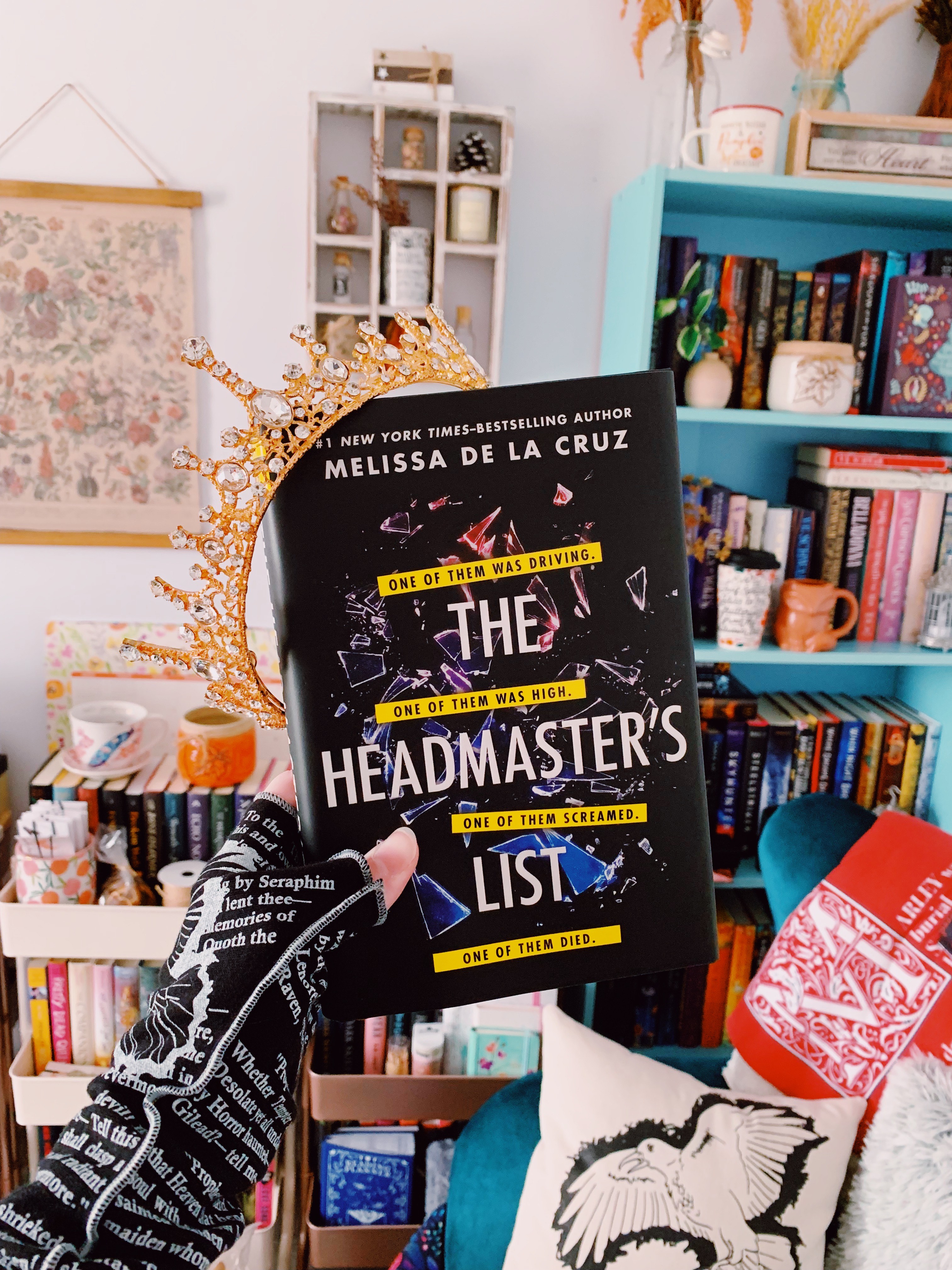 A hardcover copy of The Headmaster's List held in front of a blue bookshelf.